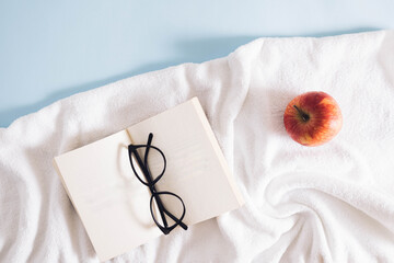 Summer scene with a bright white beach towel,  red apple, and book with glasses against a blue background. Minimal aesthetic.