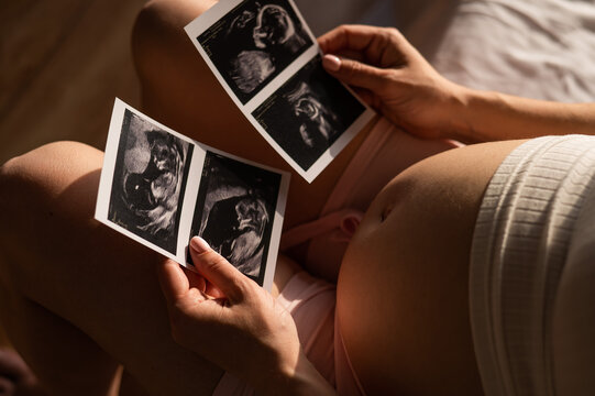 A pregnant woman looks at a photo from an ultrasound while sitting on the bed. Close-up of a pregnant woman's belly. 