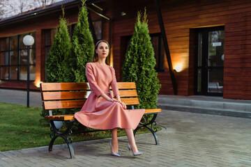 Beautiful young woman in pink dress is sitting on a wooden bench. Girl looking to the side