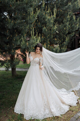 Wedding portrait. A brunette bride in a lace veil and a white dress with lace sleeves poses near a coniferous tree. The veil is in the air. Open shoulders of the bride. Photo session in nature.