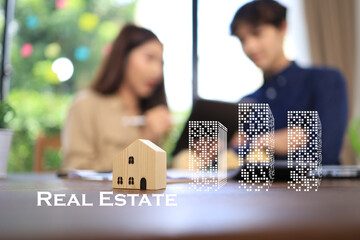 The wooden house sits on the desk of the office of a real estate company. The background is the...