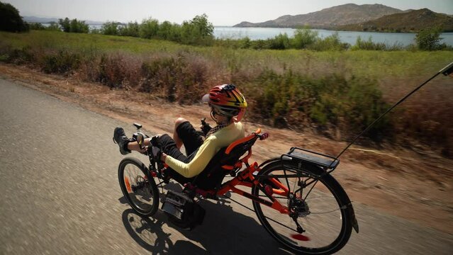 Rear view of elderly senior woman riding recumbent e-bike electric tricycle bicycle beside a lake on a sunny day beside a lake with mountains in the distance.