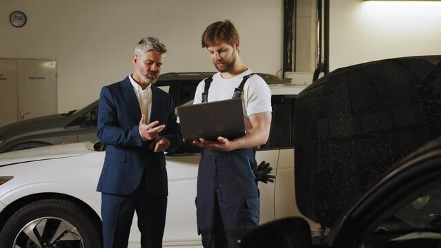 Mature grey hair business male customer with a black car and an auto mechanic looking at a laptop computer in auto-service. Vehicle wiring inspection. Car service, repair, maintenance.
