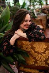 Young smiling woman with long dark hair sitting on retro sofa among plants in greenhouse