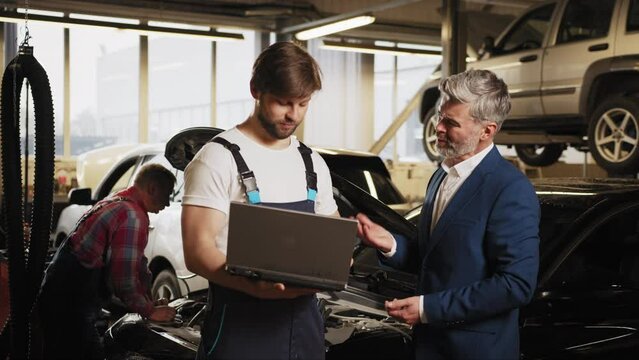 Young skilful mechanic at work in his garage with customer in auto-service. Car service, repair, maintenance and people concept. Engineer with laptop makes computer diagnostics of the car engine