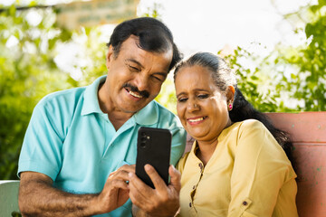 laughing Indian senior couple while watching mobile phone at park - concept of social media, technology and retirement lifestyle