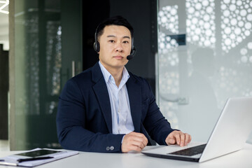 Portrait of serious businessman inside office, asian man with headset phone thinking looking at camera, man working inside office with laptop at workplace in business suit, mature boss.