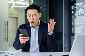 Confused Asian guy holding smart phone feels concerned thinking over received message. Mobile phone...