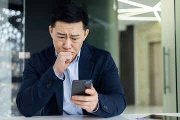 Portrait of a worried young Asian man sitting in the office at the table, holding his head with his...