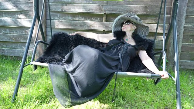 Portrait of a happy elderly woman 65 - 70 years old in a straw hat resting on a swing in the garden, close up