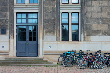 View of the facade of grey brick building, door and window with dark-blue frame and a lot of parked bicycles near. Street style. Transport in the city. Bicycle parking. Dresden, Germany, May 2023 - obrazy, fototapety, plakaty