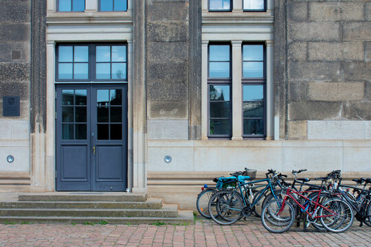 View of the facade of grey brick building, door and window with dark-blue frame and a lot of parked bicycles near. Street style. Transport in the city. Bicycle parking. Dresden, Germany, May 2023