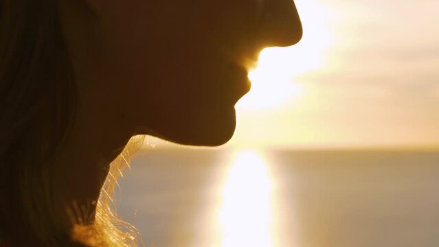 LENS FLARE, CLOSE UP: Wonderful portrait of a beautiful woman watching sunset. Golden sun's rays sparkle around her face while summer breeze ruffles her hair. She watches setting sun over Adriatic Sea