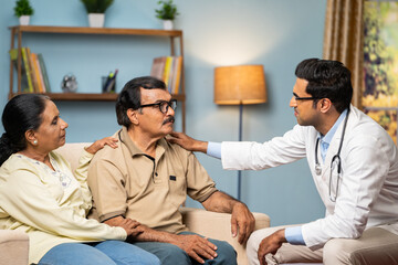 Indian doctor consoling senior couple by giving confidence while sitting on sofa at home - concept...