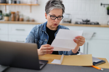 Adult woman in casual clothes surprised checks bills while sitting at table with laptop.