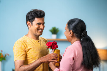 Indian adult son giving flowers with gift to senoir mother during mother day celebration - concept of relationship bonding, birthday present and happy moment