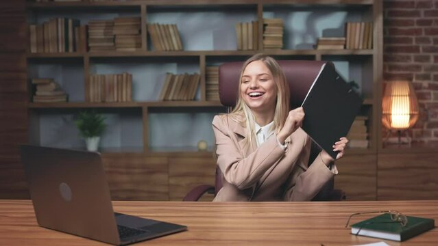 Caucasian open-minded director finishing work and dancing with financial report on comfortable office chair. Cheerful lady listening to lively music on laptop and having fun.
