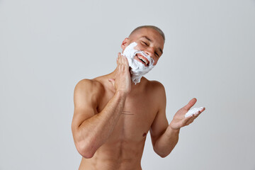 Portrait of shirtless young man applying shaving cream against white studio background. Getting ready. Concept of men's beauty, skincare, hygiene, cosmetology, health. Copy space for ad