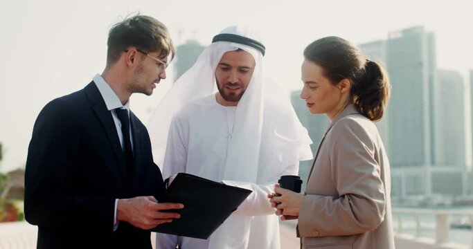A man and a woman in European business clothes, and an Arab man in a traditional dress smile while studying documents outdoors
