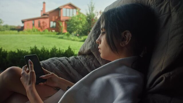 A brunette young woman is scrolling through her phone while sitting on a swing seat. There is a big house in the background. The woman has tattoos on her arm.