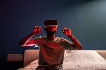 African American man wearing futuristic metaverse VR headset touching air with finger, interacting with virtual objects, shopping in virtual reality while sitting on bed under red neon light