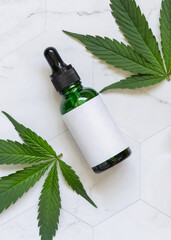 Dropper bottle with blank label near green cannabis leaves on white table. Cosmetic Mockup
