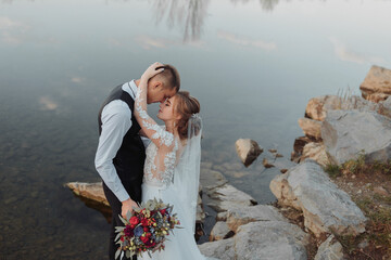 Fototapeta na wymiar brides on the bank of the river, with bare legs hugging and kissing on a rock holding a bouquet of various flowers, red roses and blue flowers and a long white dress