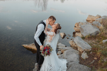 Fototapeta na wymiar brides on the bank of the river, with bare legs hugging and kissing on a rock holding a bouquet of various flowers, red roses and blue flowers and a long white dress