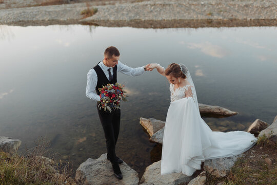 brides on the bank of the river, with bare legs walking on the rock holding a bouquet of various flowers, red roses and blue flowers and a long white dress