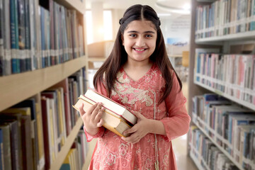 Portrait of happy smiling Indian school girl with long hair beautiful eyes wearing traditional dress costume, holds books in library, student child stands near bookshelf. Asian female kid education.