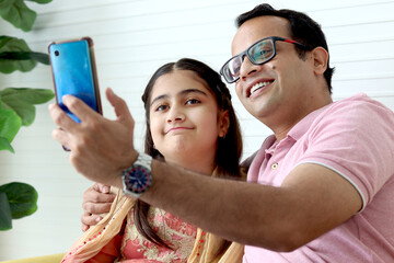 Indian happy family spending time together, cute daughter girl kid in India traditional dress costume and her father taking a selfie by mobile phone while sitting in living room or making video call.