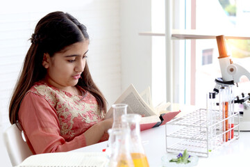 Cute Indian school girl in India traditional dress costume reading book before doing science experiments in lab, young scientist kid with microscope and lab equipment learning chemistry in classroom.