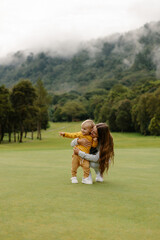 Obraz premium Happy young woman hugs and cuddles her baby while sitting on a green lawn with a beautiful view of the nature garden. Family outdoor recreation, happy family