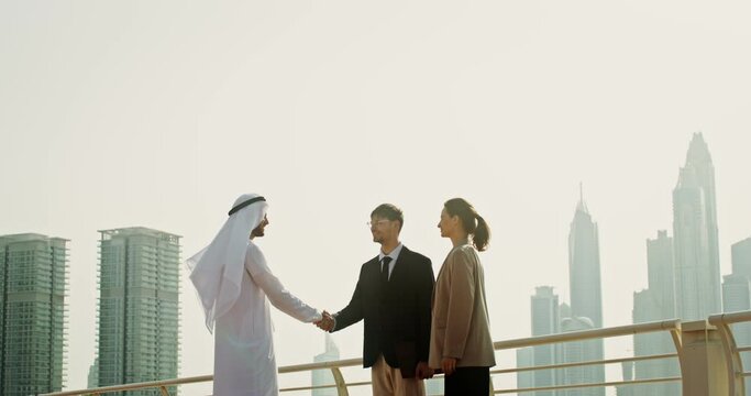 A young man in Arabic traditional white clothes shakes hands with his European colleagues while standing outdoors