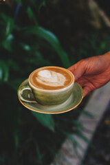 Hand holding a mug of cappuccino against the background of plants