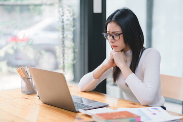 The stressed and exhausted millennial Asian businesswoman is seen sitting at her office desk with...