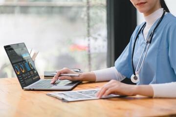 A professional female doctor is taking notes in a medical journal and filling out documents, including the patient's illness history, looking at a laptop screen dashboard.