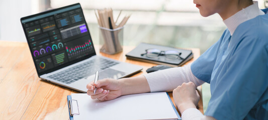 Cropped shot of a professional female doctor is taking notes in a medical journal and filling out documents, including the patient's illness history, looking at a laptop screen dashboard.