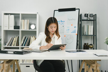 A portrait of a smiling, young, beautiful, professional, and confident millennial Asian businesswoman using a digital tablet to analyze sales data at office.