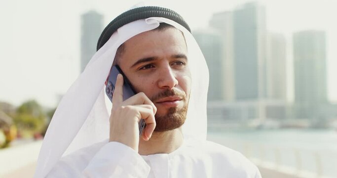 A young man in Arab national white clothes smiles while talking on a mobile phone while standing in the open air. Face close-up