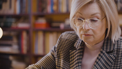 Bored senior woman reading historical books in a library late at night, student