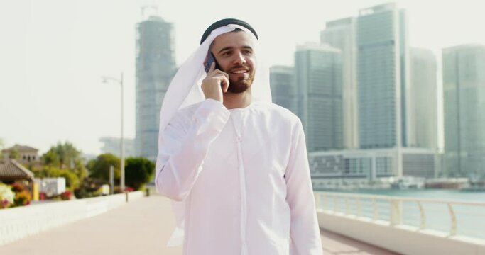 A young man in Arab national white clothes smiles, while talking on the phone walking along the embankment of a modern city on a sunny day - Powered by Adobe