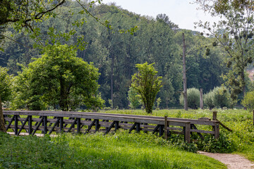 Fototapeta premium Valley with a wooden bridge over Geul river in Dutch nature park Ingendael, mountain covered with lush trees in background, sunny spring day on hiking trail, South Limburg in the Netherlands