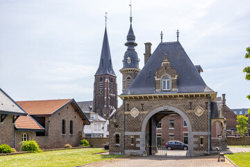 Back of arched entrance to Borgharen castle against blue sky, dome with small window, circular tower, brick walls and fence, church steeple in background, sunny day in South Limburg, Netherlands