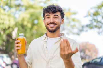 Young Arabian handsome man holding an orange juice at outdoors inviting to come with hand. Happy...