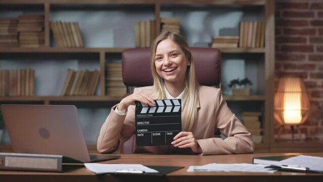 Front view of elegant young woman holding black movie clapper in hands and smiling at camera in studio. Vibrant lady dressed in trendy beige jacket participating in film industry.