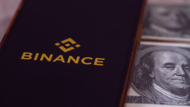 Saraburi, Thailand - 06-11-2023: On  screen mobile have logo of the binance trading website. And there are bnb coins and dollars laying nearby.