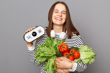 Time top eat fresh food. Vegetarian diet promotes health. Healthy lifestyle with green choices. Smiling woman holding vegetables and showing clock isolated over gray background.