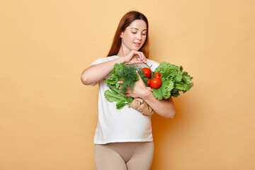 Future mother buying organic food, pregnant woman holding fresh vegetables posing isolated over beige background, prefers useful balanced diet.