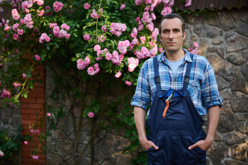 Portrait of a handsome Caucasian young man, gardener in gardening outfit, looking at camera, standing at his workplace in the backyard of a mansion, over background of blooming rose